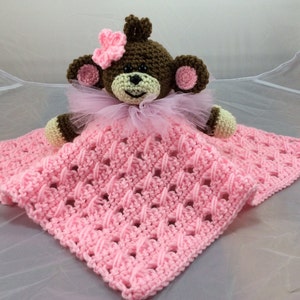 May include: A pink crocheted baby blanket with a brown crocheted monkey toy on top. The monkey has a pink flower on its head and a pink tulle bow around its neck.