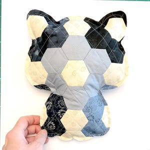 Può includere: Un cuscino fatto a mano a forma di gatto con un design patchwork. Il cuscino presenta una varietà di tessuti in crema, grigio e blu navy, cuciti insieme in un motivo esagonale. Le orecchie sono blu navy scuro e il cuscino è trapuntato con filo bianco.