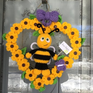 Puede incluir: Una corona de abeja de crochet con flores amarillas, hojas verdes y un lazo morado. La abeja sostiene un cartel que dice "Bee Happy".