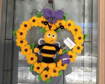 Könnte beinhalten: Ein gehäkelter Bienen-Kranz mit gelben Blumen, grünen Blättern und einer lila Schleife. Die Biene hält ein Schild mit der Aufschrift "Bee Happy".