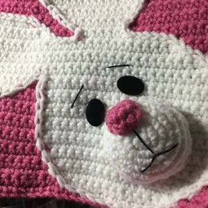 May include: A crocheted pink and white bunny blanket with black button eyes and a pink nose.