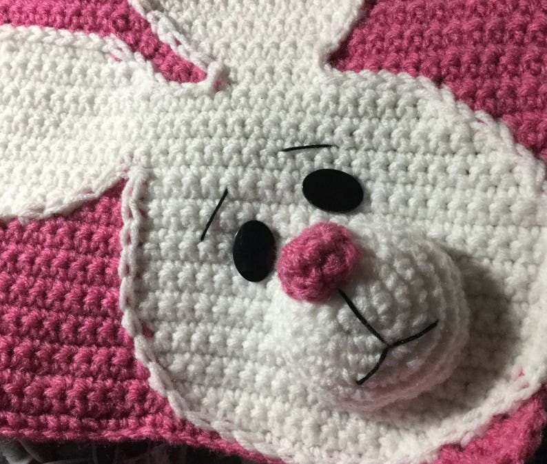 May include: A crocheted pink and white bunny blanket with black button eyes and a pink nose.