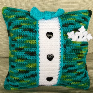May include: A crocheted pillow with a tuxedo design. The pillow is green, blue, and white with black heart-shaped buttons. The pillow has a pocket on the side with white crocheted snowflakes.