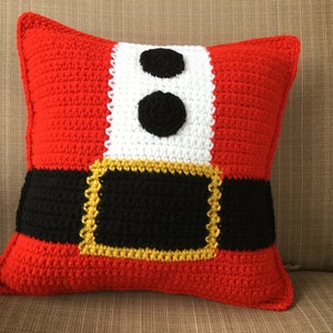May include: A red, white, and black crocheted pillow with a belt and two black buttons. The pillow is shaped like a Santa Claus suit.
