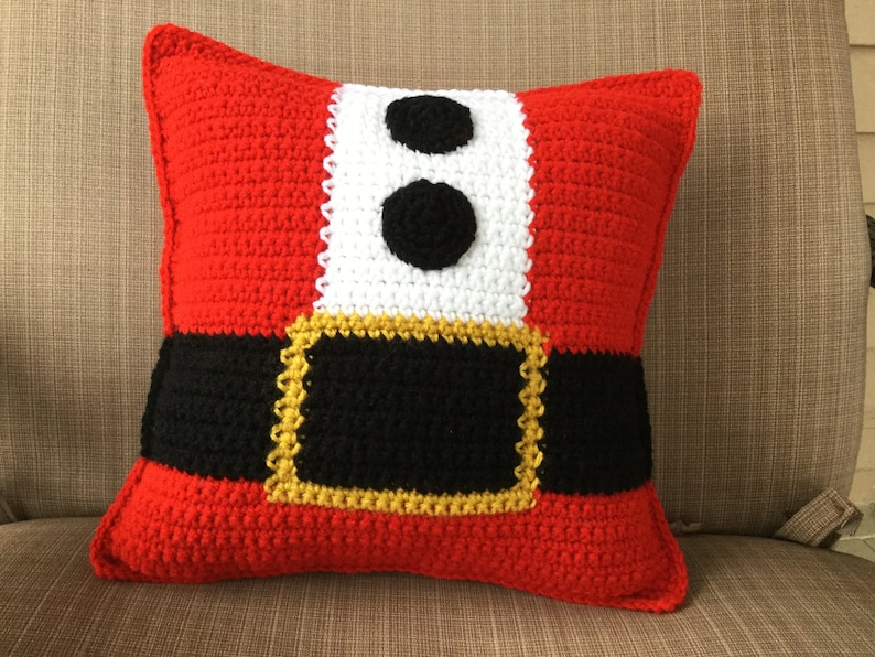 May include: A red, white, and black crocheted pillow with a belt and two black buttons. The pillow is shaped like a Santa Claus suit.
