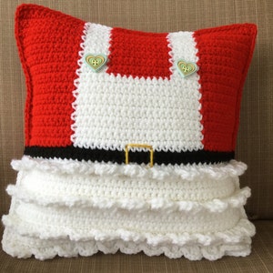 May include: A red and white crocheted pillow with a black belt and white ruffles. The pillow has two green heart-shaped buttons.
