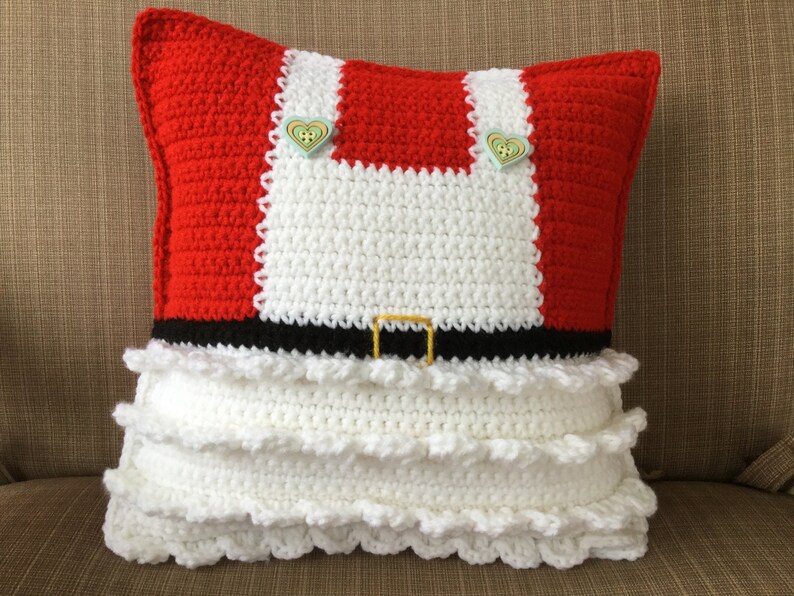 May include: A red and white crocheted pillow with a black belt and white ruffles. The pillow has two green heart-shaped buttons.