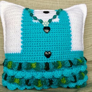 May include: A crocheted pillow in the shape of a dress with a turquoise bodice, white sleeves, and a green and turquoise ruffled skirt. The pillow has a turquoise beaded necklace with a snowflake charm and two black heart-shaped buttons.