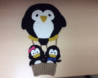 Könnte beinhalten: Eine handgefertigte Häkelarbeit mit einer Pinguinfamilie in einem Heißluftballon-Design. Der Ballon ist schwarz und weiß, mit gelbem Schnabel und Füßen. Zwei kleinere Pinguine befinden sich in einem braunen Korb, einer mit rosa Schleife, der andere mit blauer Fliege.