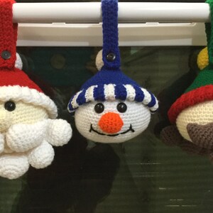 May include: Three crocheted Christmas ornaments hanging from a white bar. The ornaments are a snowman, Santa Claus, and an elf. The snowman is wearing a blue and white striped hat, the Santa Claus is wearing a red hat with white trim, and the elf is wearing a green hat with a red and white trim.