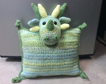May include: A green and yellow crocheted dragon pillow with a cute face. The pillow is made with a soft, textured yarn and has a striped pattern.
