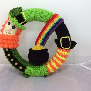 May include: A crocheted wreath featuring a leprechaun, a pot of gold, and a rainbow. The leprechaun is wearing a green hat and has a red beard. The pot of gold is black with a yellow lid. The rainbow is multi-coloured.