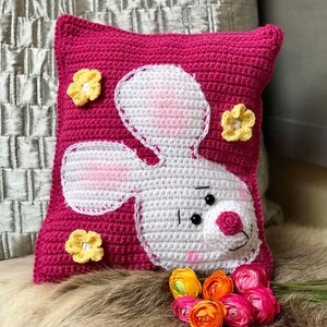 May include: A pink crocheted pillow with a white crocheted bunny face and yellow crocheted flowers.
