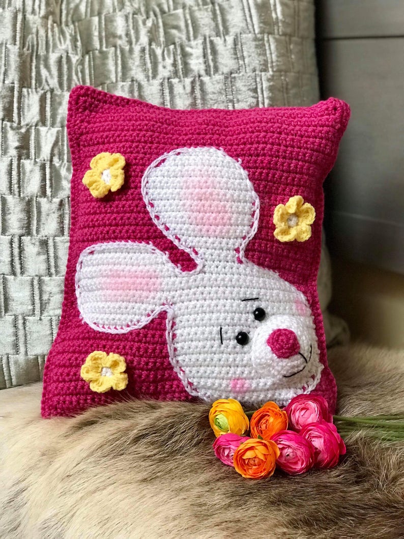 May include: A pink crocheted pillow with a white crocheted bunny face and yellow crocheted flowers.