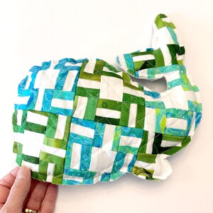 May include: A handmade whale-shaped fabric item with a patchwork design. The whale features a combination of white, green, and blue fabric pieces, stitched together. The item is quilted and has a three-dimensional shape, with a tail and a head.