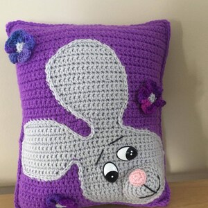 May include: Purple crocheted pillow with a gray crocheted bunny face and purple crocheted flowers.