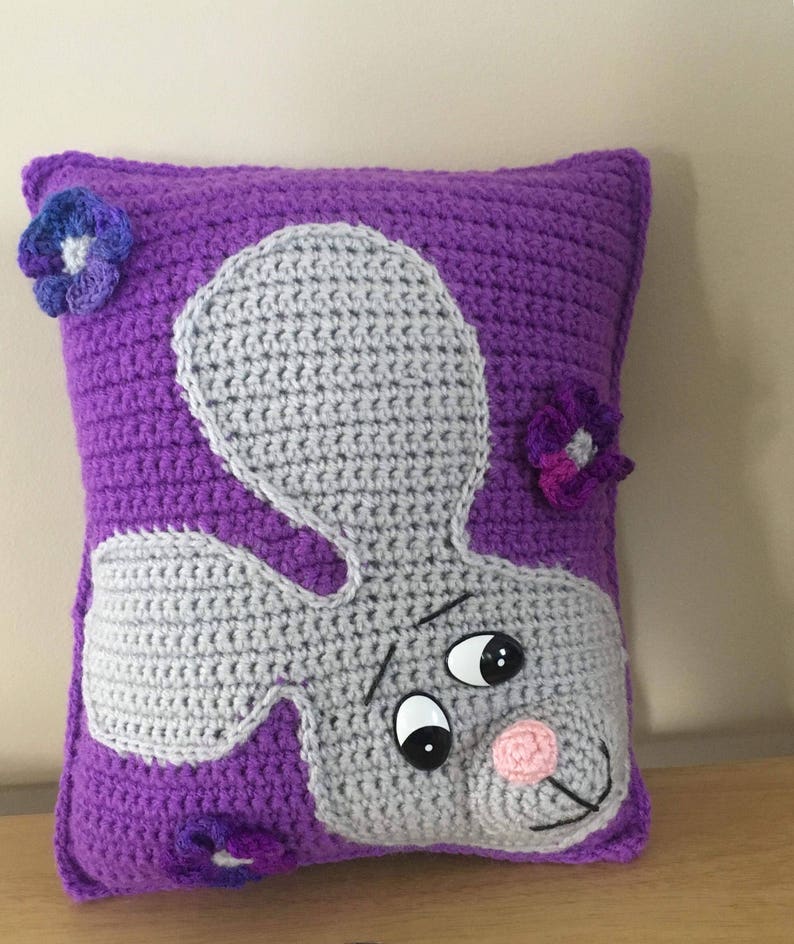 May include: Purple crocheted pillow with a gray crocheted bunny face and purple crocheted flowers.