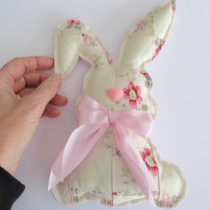 May include: A fabric bunny with a pink bow and heart-shaped nose. The bunny is cream-coloured with a floral pattern in pink and green. The bunny's ears are upright, and a hand is holding the ear.