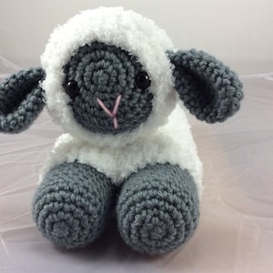Op de afbeelding: Een gehaakt knuffellammetje met een wit, pluche lijf en grijze gehaakte poten, oren en gezicht. Het lammetje heeft zwarte ogen en een roze neus.