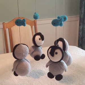 May include: A crocheted baby mobile with gray and black penguins and blue fish hanging from a gray crocheted ring.