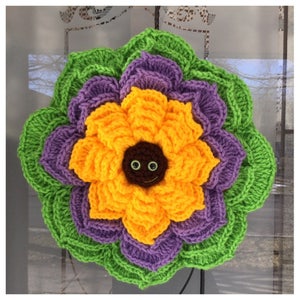 May include: A crocheted sunflower with a brown center and googly eyes. The sunflower is surrounded by layers of purple and green crocheted petals.