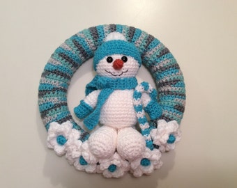 May include: A handmade crochet snowman wreath. The wreath is a ring of blue, grey, and white yarn. A white snowman with a blue hat, scarf, and mittens is in the centre. White flowers with blue centres decorate the bottom.