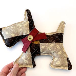 May include: A handmade fabric dog-shaped pillow. The pillow is made of patchwork fabric in shades of brown and beige with a leaf pattern. It has a burgundy bow with a button and is held by a hand.