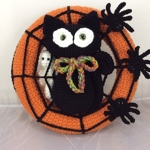 May include: A black crocheted cat with green and purple yarn bow sits in an orange crocheted spiderweb wreath. Three black crocheted spiders are attached to the wreath.