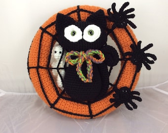 May include: A black crocheted cat with green and purple yarn bow sits in an orange crocheted spiderweb wreath. Three black crocheted spiders are attached to the wreath.