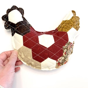 May include: A quilted chicken-shaped potholder in burgundy, brown, cream, and gold. The patchwork design features geometric shapes and diamond quilting. The chicken has a ruffled comb and wattle.