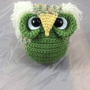 May include: A crocheted owl toy made with green, brown and white yarn. The owl has large, white eyes with black pupils and a yellow beak.