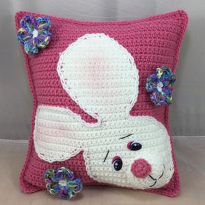 May include: Pink crocheted pillow with a white bunny face and colorful flower embellishments.