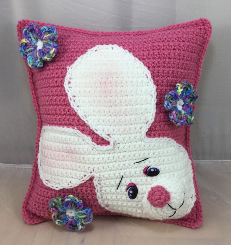 May include: Pink crocheted pillow with a white bunny face and colorful flower embellishments.