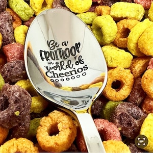 May include: A silver spoon with a gold handle, engraved with the text "Be a Fruitloop in a world of Cheerios". The spoon is resting on a bed of colourful cereal.