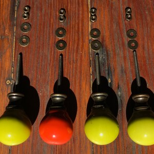 以下が含まれることがあります： 金属製のベースとネジ付きボルトを備えた4つの丸いキャビネットノブ。3つのノブは明るい黄色で、1つはオレンジ色です。各ノブには2つの金属ワッシャーと2つのナットが含まれており、対応するノブのハードウェアの上に垂直に並べられています。