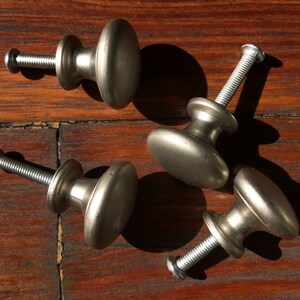 Pode incluir: Quatro puxadores de armário em metal prateado com um topo liso e arredondado e uma base cónica. Cada puxador possui um parafuso de metal roscado que se estende a partir da parte de trás, concebido para fixação em móveis ou portas.