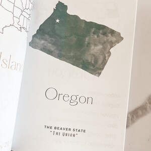 May include: Open book page featuring a watercolor-style map of Oregon in shades of green and gray. The word "Oregon" is printed in a minimalist font, with the state's nickname, "The Beaver State," below.