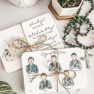 Puede incluir: Dos tarjetas con ilustraciones y texto. La primera tarjeta muestra un dibujo de una persona con alas y un halo. El texto en la tarjeta dice "&iquest;Qui&eacute;n te hizo? Dios me hizo. &iquest;Dios hizo todas las cosas? S&iacute;. Dios hizo todas las cosas. &iquest;A qui&eacute;n hizo Dios? Dios me hizo para mostrar su y hacerme feliz con". La segunda tarjeta muestra una serie de ilustraciones de una persona haciendo la se&ntilde;al de la cruz. El texto en la tarjeta dice "Se&ntilde;al de la cruz. En el nombre del Padre. Y del Hijo. Y del Esp&iacute;ritu Santo. Am&eacute;n."