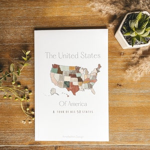 May include: A white book titled "The United States Of America" with a watercolor map of the U.S. and the text "A tour of all 50 states." The book is on a wooden surface with a small succulent plant.