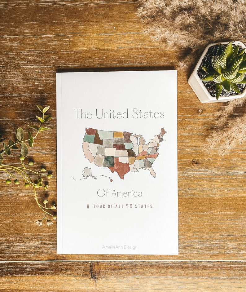 May include: A white book titled "The United States Of America" with a watercolor map of the U.S. and the text "A tour of all 50 states." The book is on a wooden surface with a small succulent plant.