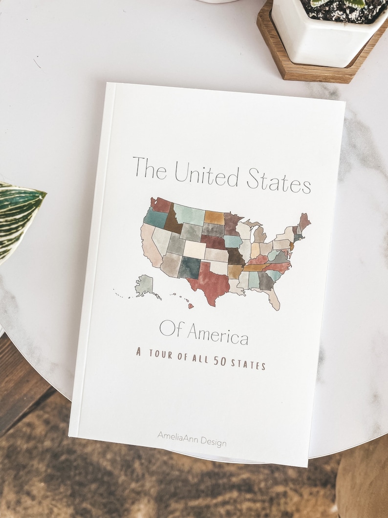 May include: A white book titled "The United States Of America" with a watercolor map of the U.S. and the text "A TOUR OF ALL 50 STATES". The book is on a white marble table.