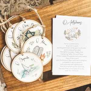May include: A set of circular wooden decorations with watercolour illustrations and text, strung with twine. The ornaments depict a dove, a star, and other religious symbols. A card with text is also present.