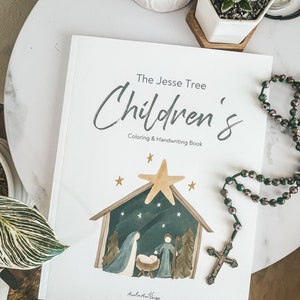 Pode incluir: Um livro branco para colorir e escrever para crianças intitulado "The Jesse Tree" com uma ilustração de uma cena de presépio. Um rosário com contas verdes e uma cruz está ao lado do livro. O livro está numa mesa branca com plantas.