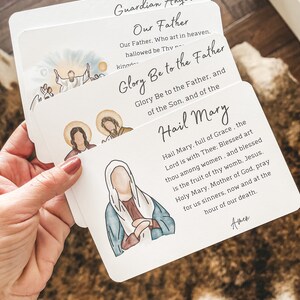 Puede incluir: Conjunto de cuatro tarjetas de oraci&oacute;n blancas con ilustraciones de figuras religiosas. Las tarjetas presentan las oraciones "&Aacute;ngel de la Guarda", "Padre Nuestro", "Gloria al Padre" y "Ave Mar&iacute;a".