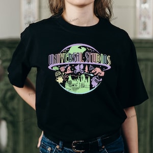 May include: Black t-shirt featuring a colorful graphic of the Universal Studios logo with various cartoon characters and a castle silhouette. The text "UNIVERSAL STUDIOS" is arched above the graphic.