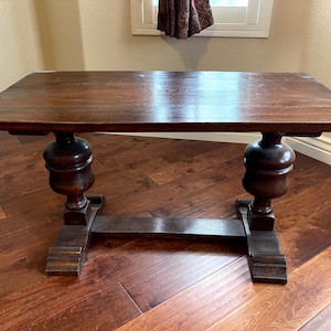 May include: A rectangular dark brown wooden table featuring a smooth flat top surface supported by two thick, bulbous pedestal legs connected by a horizontal base stretcher that rests on two wide, flat feet on each side of the furniture piece.