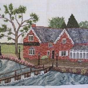 May include: Cross-stitch of a brick building with a gray roof, labeled "The Haven". A bridge crosses a body of water in the foreground, with a tree and flowers on the left. The scene is detailed with various colors.