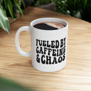 Puede incluir: Taza de café de cerámica blanca con un gráfico negro que dice "Fueled by Caffeine & Chaos".