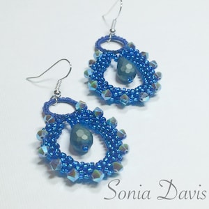 May include: A pair of handmade blue beaded earrings. Each earring features a teardrop-shaped design with a central blue gemstone and a ring at the top. The earrings are adorned with iridescent beads and have silver-colored ear wires. The name "Sonia Davis" is in the bottom right corner.