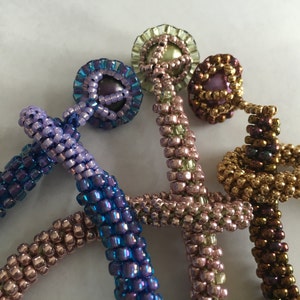 May include: Close-up of three beaded bottle stoppers in shades of purple, gold, and pink. Each stopper features a beaded stem and a decorative beaded top. The beads have a metallic sheen, adding a touch of elegance.